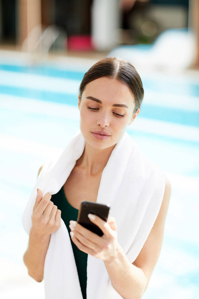 Νεαρή γυναίκα με μεγάλη λευκή μαλακή πετσέτα γραπτών μηνυμάτων σε smartphone στις διακοπές - Φωτογραφία, εικόνα