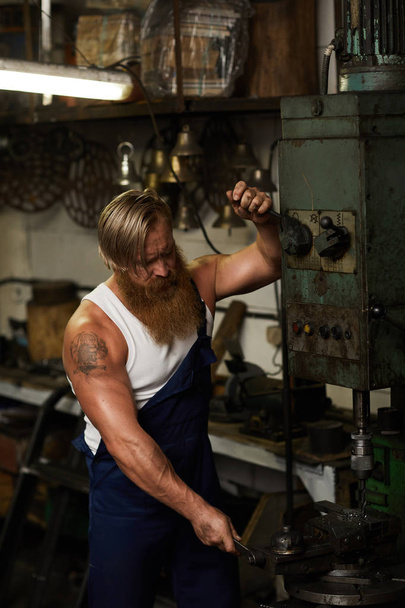 Artisan musclé concentré sérieux avec tatouage et longue barbe travaillant avec une machine de forage et utilisant des poignées pour fixer les détails
 - Photo, image