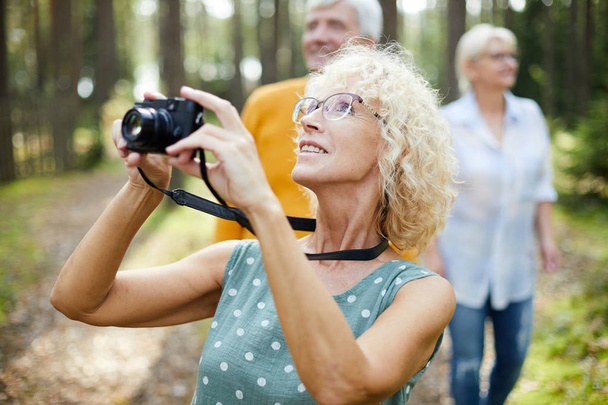 Smiling excited mature lady with curly hair wearing glasses looking up and photographing animal in forest while walking with friends - Photo, image