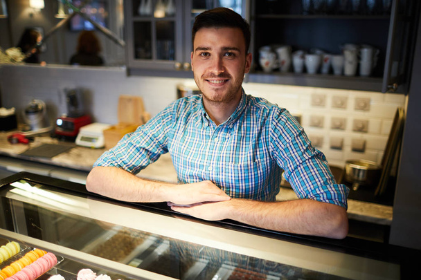 Młody sprzedawca patrząc w aparat przy dużym wyświetlaczu z deserami i ciastem w kawiarni - Zdjęcie, obraz