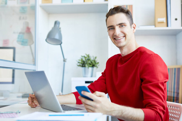 Усміхнений молодий чоловік у повсякденному одязі використовує сучасні гаджети, сидячи за столом в офісі
 - Фото, зображення