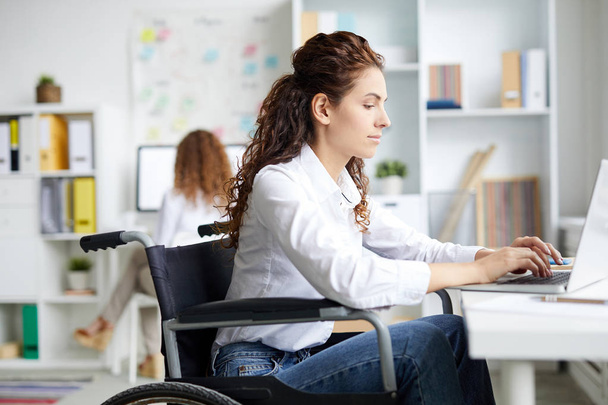 Młoda bizneswoman siedzi na wózku inwalidzkim i pisze na klawiaturze laptopa w biurze - Zdjęcie, obraz