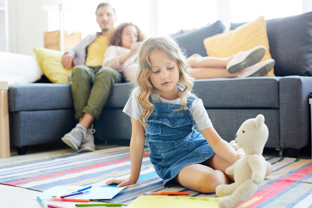 Cute little daughter playing with teddybear on the floor with her parents relaxing on sofa - Foto, imagen