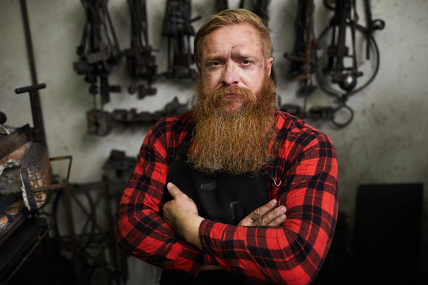 Portrait of serious confident handsome male smith with long beard looking scowling and crossing arms on chest in workshop - Photo, image