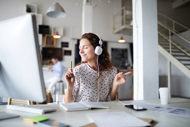Joyous young businesswoman with headphones listening to dynamic music while working by desk in front of computer monitor - Foto, Imagem