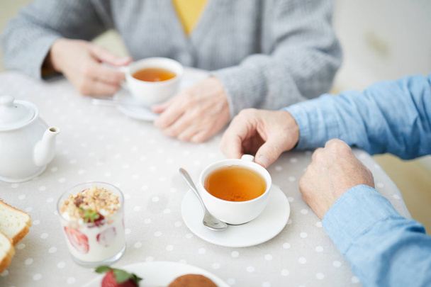 Руки пожилой пары за столом с чашками напитков, клубничный десерт в стекле и белый фарфоровый чайник во время чая
 - Фото, изображение