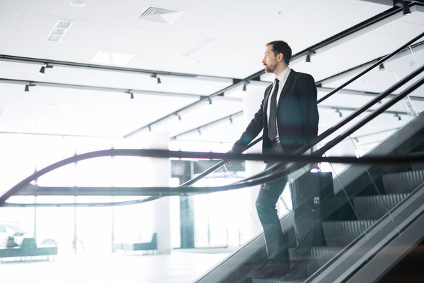 Young confident businessman with briefcase standing on steps of escalator and moving downwards - Foto, Imagem