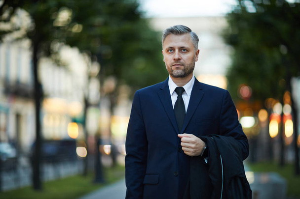 reifer seriöser Geschäftsmann im eleganten Anzug, der seinen Mantel hält, während er in der städtischen Umgebung spaziert - Foto, Bild