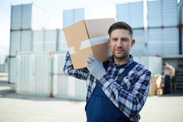 Νεαρός άνδρας με μεγάλο κουτί κοιτάζει την κάμερα ενώ μεταφέρουν φορτίο στην αποθήκη - Φωτογραφία, εικόνα