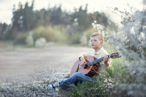 Outdoor portrait of a young teenager playing guitar and sitting on the road near park. - Zdjęcie, obraz