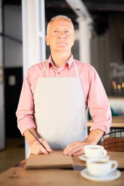 Старший официант или владелец кафе в рабочей одежде встречи гостей и делать рабочие заметки в блокноте
 - Фото, изображение