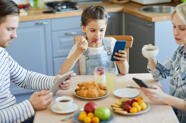 Nowoczesna rodzina za pomocą mobilnych gadżety, podczas gdy śniadanie serwowane przez stół w kuchni - Zdjęcie, obraz