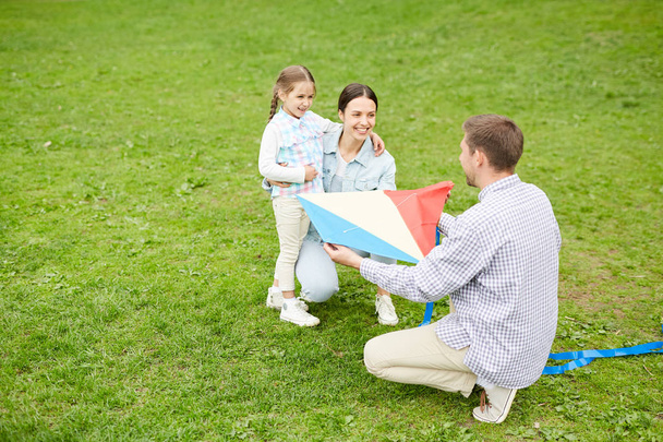 Jongeman tonen zelfgemaakte vlieger aan zijn vrouw en dochtertje tijdens het spelen op groen gazon - Foto, afbeelding