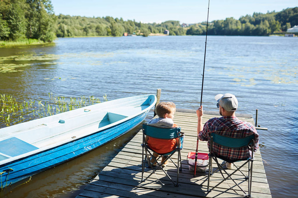 Vater und Sohn sitzen auf Stühlen auf der Seebrücke und angeln gemeinsam in der schönen Natur - Foto, Bild