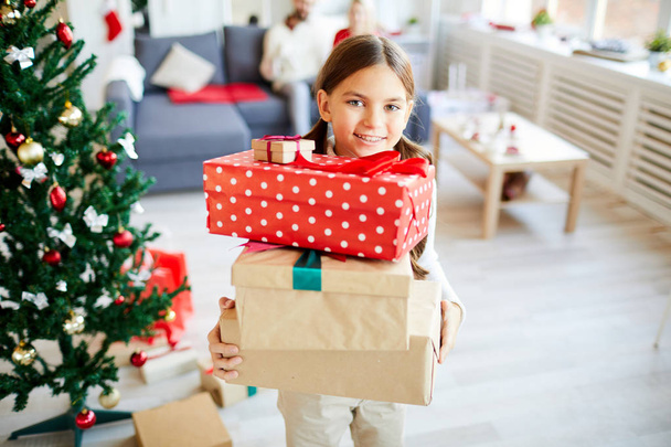 Little girl showing you stack of Christmas gifts while standing by decorated firtree - Photo, Image