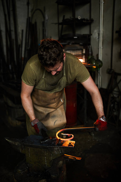 Молодой кузнец в рабочей одежде наклоняется над наковальней и работает с молотком и расплавленным металлом заготовки
 - Фото, изображение
