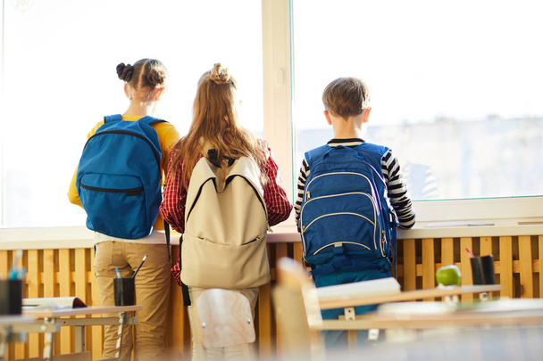 Πίσω όψη του ονειρεμένο σχολείο φίλων με χαρτοφύλακες σε πλάτες κοιτάζοντας έξω παράθυρο, ενώ περιμένουν για τους γονείς στην τάξη - Φωτογραφία, εικόνα