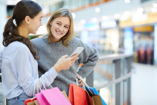 Uno de los jóvenes compradores mirando a su amiga mostrando curiosa notificación de venta en el teléfono inteligente después de comprar en el centro comercial
 - Foto, Imagen
