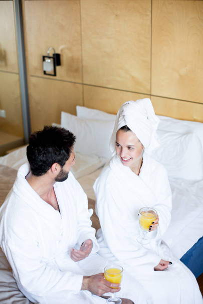 Szczęśliwy młody para w szlafroki siedzący na łóżko w Hotel pokój, rozmowa i mający orzeźwiający napój - Zdjęcie, obraz