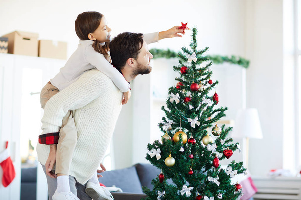 Little girl sitting on father back while putting red decorative star on top of Christmas tree - Φωτογραφία, εικόνα