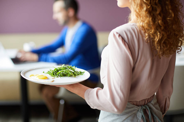 Закрыть официантку в розовой блузке, несущую жареные яйца с зеленью посетителю ресторана во время завтрака
 - Фото, изображение
