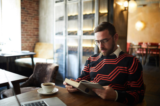 Спокійний чоловік у повсякденному светрі сидить за столом у кафе, читає книгу та п'є на дозвіллі
 - Фото, зображення