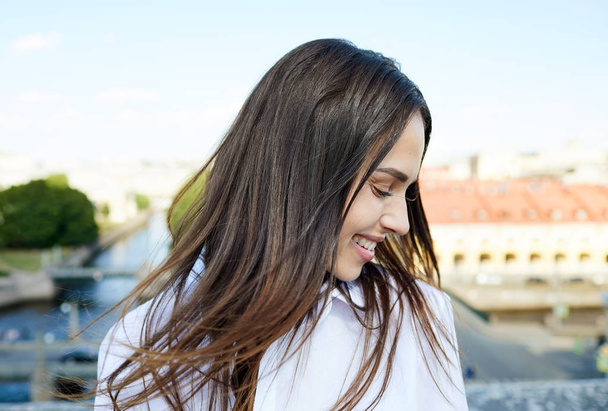 Ευτυχισμένο όμορφο κορίτσι με μαύρα μαλλιά κρατώντας τα μάτια κλειστά, ενώ νιώθοντας άνεμο και απολαμβάνοντας τον καιρό στην πόλη - Φωτογραφία, εικόνα
