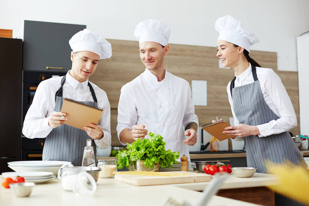 Content professional chef in white uniform giving lecture at cooking class and checking notes of students while they standing at kitchen counter with food - Photo, Image