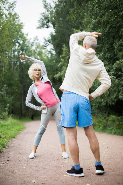 Aktywna dojrzała para w odzieży sportowej wykonuje ćwiczenia fizyczne w parku wśród zielonych drzew - Zdjęcie, obraz
