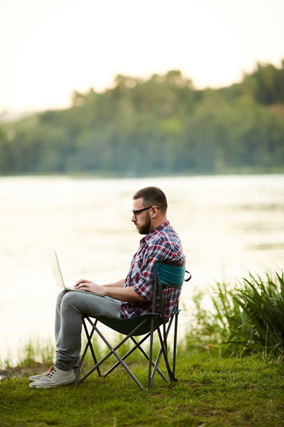 Active and mobile young man with laptop sitting by riverside and networking in the country - Foto, Imagen