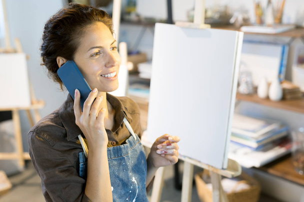 Feliz joven pintor hablando por teléfono inteligente en el taller mientras toma un descanso
 - Foto, Imagen