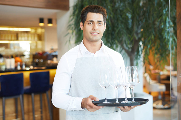 Porträt eines jungen Kellners in Uniform, der Tablett mit leeren Weingläsern im Restaurant hält - Foto, Bild