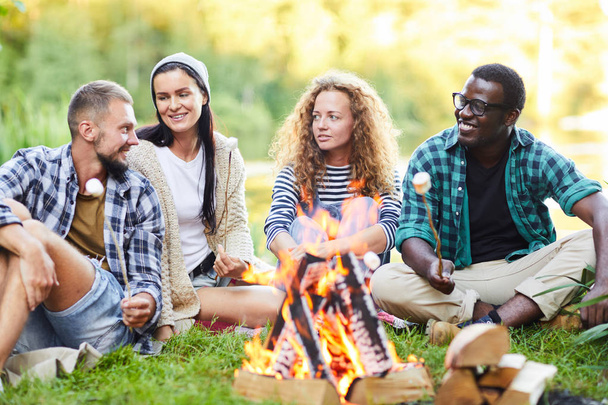 Grupa młodych obozowiczów siedzących przy ognisku i smażenia marshmellows podczas rozmowy ze sobą - Zdjęcie, obraz