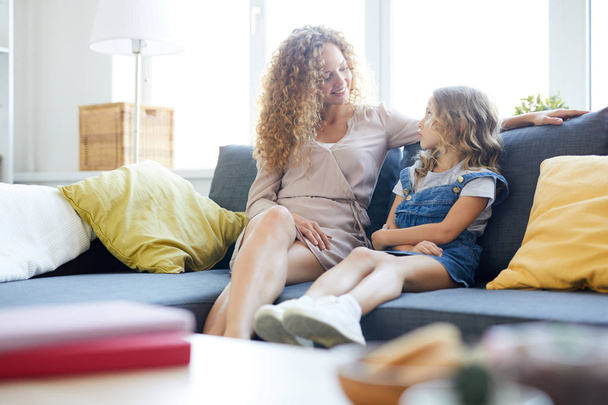 Young mother and her little daughter sitting on sofa at home, relaxing and chatting - Foto, Bild
