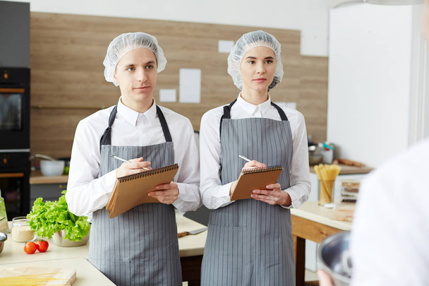 ernsthafte konzentrierte Köche in sterilen Mützen und entkleideten Schürzen, die in Notizblock schreiben, während sie dem Koch beim Kochworkshop aufmerksam zuhören - Foto, Bild