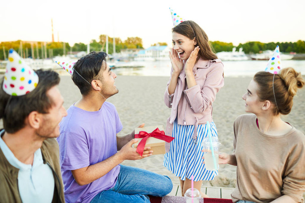 Здивована дівчина дивиться на свого хлопця з подарунковою коробкою на вечірці на пляжі
 - Фото, зображення