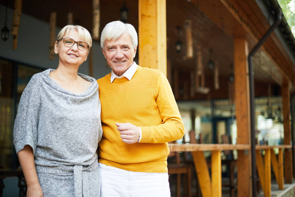 Pozitivní, šedovlasý starší muž ve žlutém svetru s milovanou přítelkyní oblečenou do šedých šatů, stojí na verandě a usmívají se na kameru - Fotografie, Obrázek