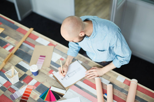 Σοβαρός άντρας δημιουργικός σχεδιαστής μόδας σε μπλε πουκάμισο στέκεται στο τραπέζι με τα δείγματα χρωμάτων και τα χαρτιά και κάνοντας νέο σκίτσο του φορέματος στο στούντιο - Φωτογραφία, εικόνα