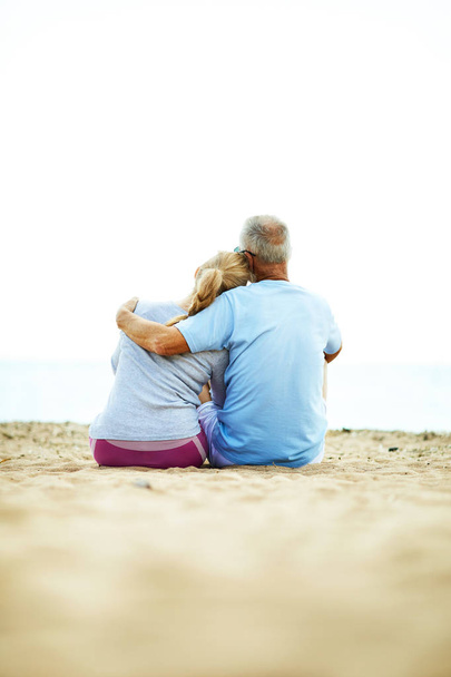 Rückansicht eines liebevollen Senioren-Paares, das nach dem Training in Umarmung auf Sand sitzt und sich entspannt - Foto, Bild