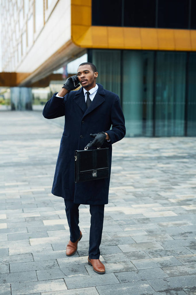 junger afrikanisch-amerikanischer Geschäftsmann ruft Kollegen oder Kunden an, während er sich im Freien bewegt - Foto, Bild
