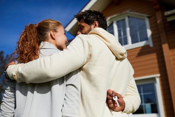 Joven abrazando a su esposa mientras sostiene la llave de la nueva casa detrás de la espalda
 - Foto, imagen
