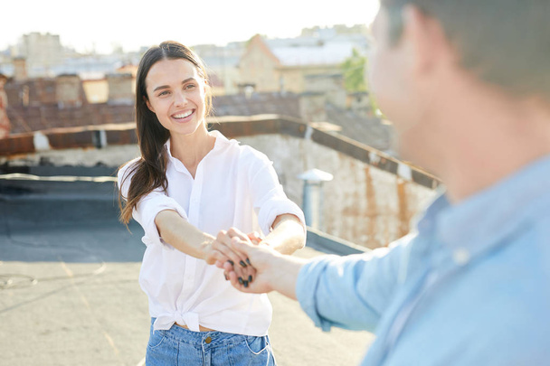 Cheerful excited pretty girl in white shirt giving hands to boyfriend while they walking boyfriend on roof at romantic date - Foto, imagen