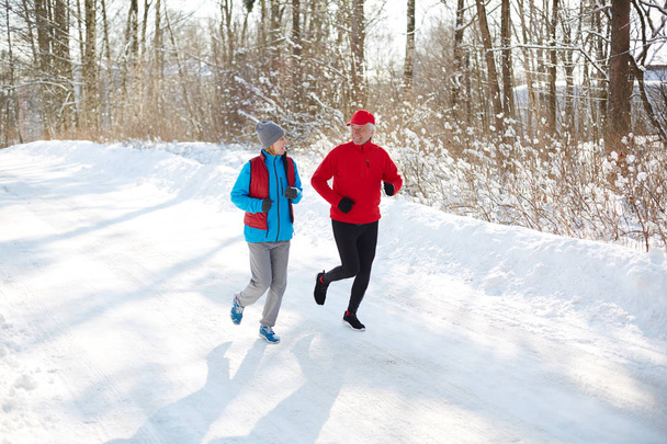 Pareja deportiva envejecida conversando mientras corre por la carretera de nieve en el soleado día de invierno
 - Foto, imagen