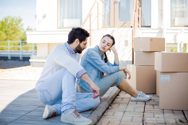 Νεαρό ζευγάρι σε ανεπίσημο ντύσιμο κάθονται στο έδαφος με στοίβα από συσκευασμένα κουτιά και το νέο τους σπίτι - Φωτογραφία, εικόνα