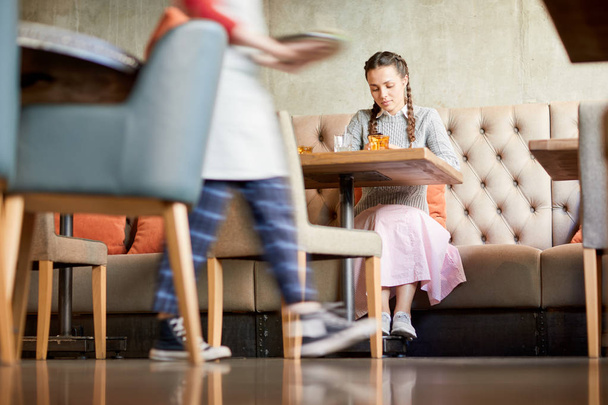 Mooi meisje zittend op de Bank in café en scrollen in smartphone tijdens het wachten op haar bestelling - Foto, afbeelding