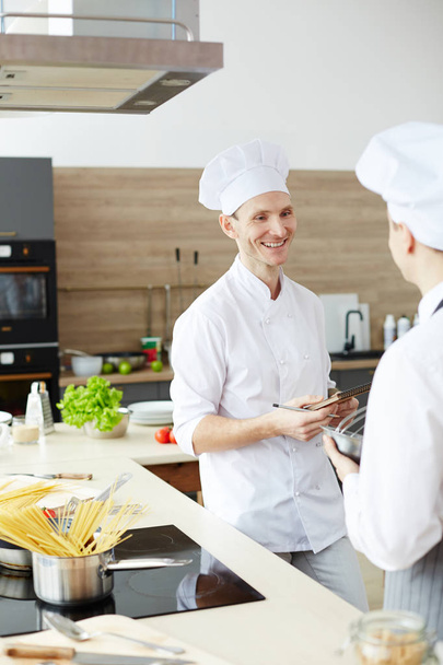 Θετικός όμορφος σεφ με στολή με στυλό και σημειωματάριο διδασκαλία νέος μάγειρας και χαμογελαστός κατά τη διάρκεια της επικοινωνίας στην εμπορική κουζίνα - Φωτογραφία, εικόνα