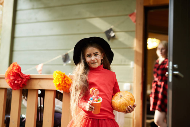 Little girl in red halloween attire and black hat holding paper glass with sweet treats and ripe pumpkin - Photo, Image