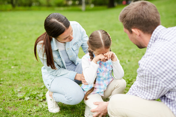 草の上に座っている間に泣いている小さな女の子と彼女の両親は週末の寒さの間に彼女を慰める - 写真・画像