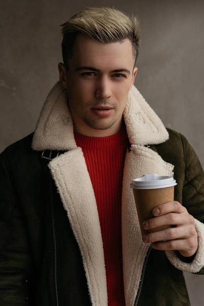 Young attractive man drinking coffee in cafe. Portrait of positive hipster guy with stylish haircut holding cup of drinks, standing near grey background and posing for photo. Coffee break concept. - Foto, Bild