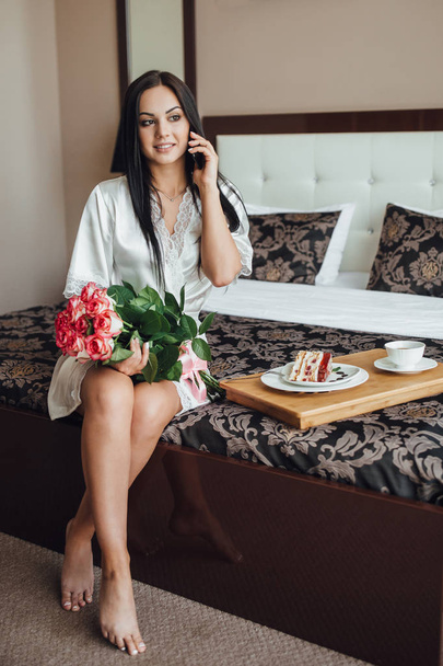 beautiful brunette woman sitting on bed in room, talking on phone and holding bunch of roses  - Foto, afbeelding